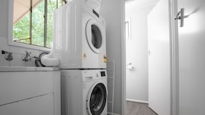 A laundry room with two stacked front-loading washing machines and a dryer, a window, and a door leading to another room. - Sweethome2br@highton+parking (Highton)