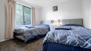 A bedroom with two beds, a window with curtains, a bedside table with lamps, and a wall art piece. - Sweethome2br@highton+parking (Highton)