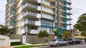 A multi-story residential building with balconies, a parking area, and a clear sky.
