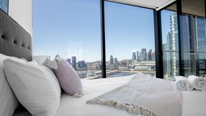 A modern hotel room with a large bed, white bedding, and a view of a city skyline through floor-to-ceiling windows. - Central Melbourne 2br2bt APT on Collins St+parking (Docklands)