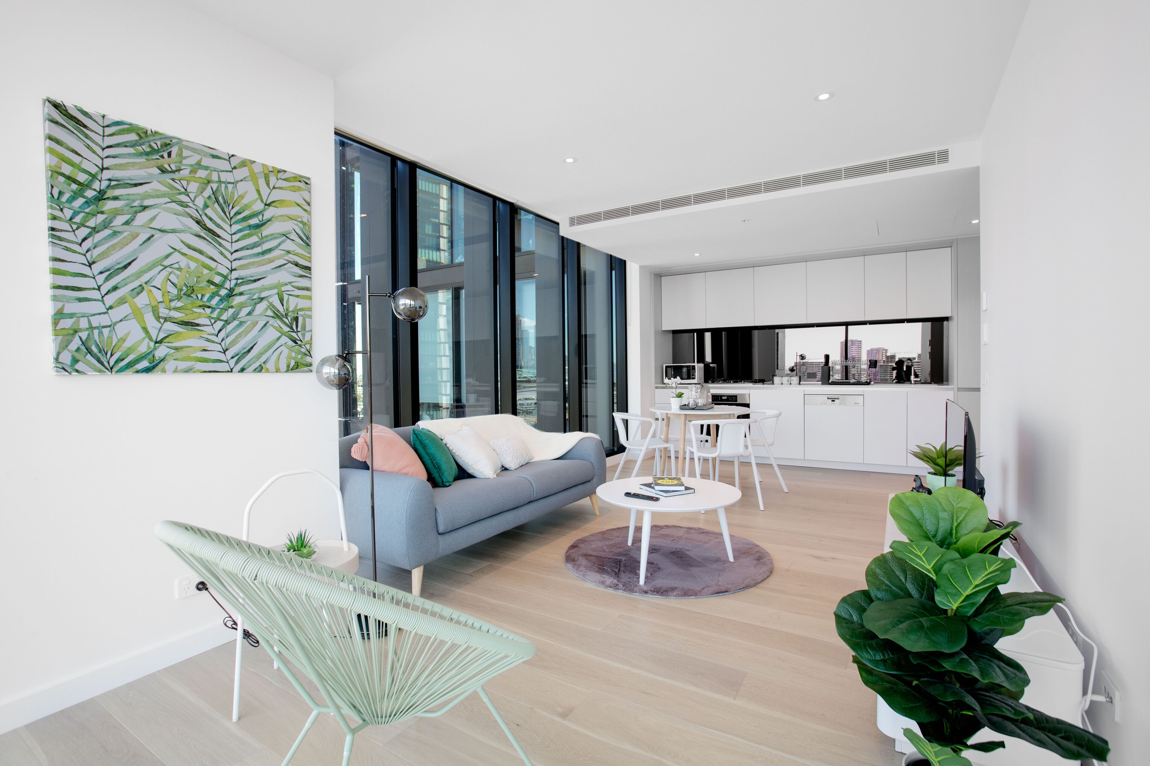 A modern living room with a sofa, a dining table, and chairs, a kitchen area with white cabinets, and a large window offering a city view.