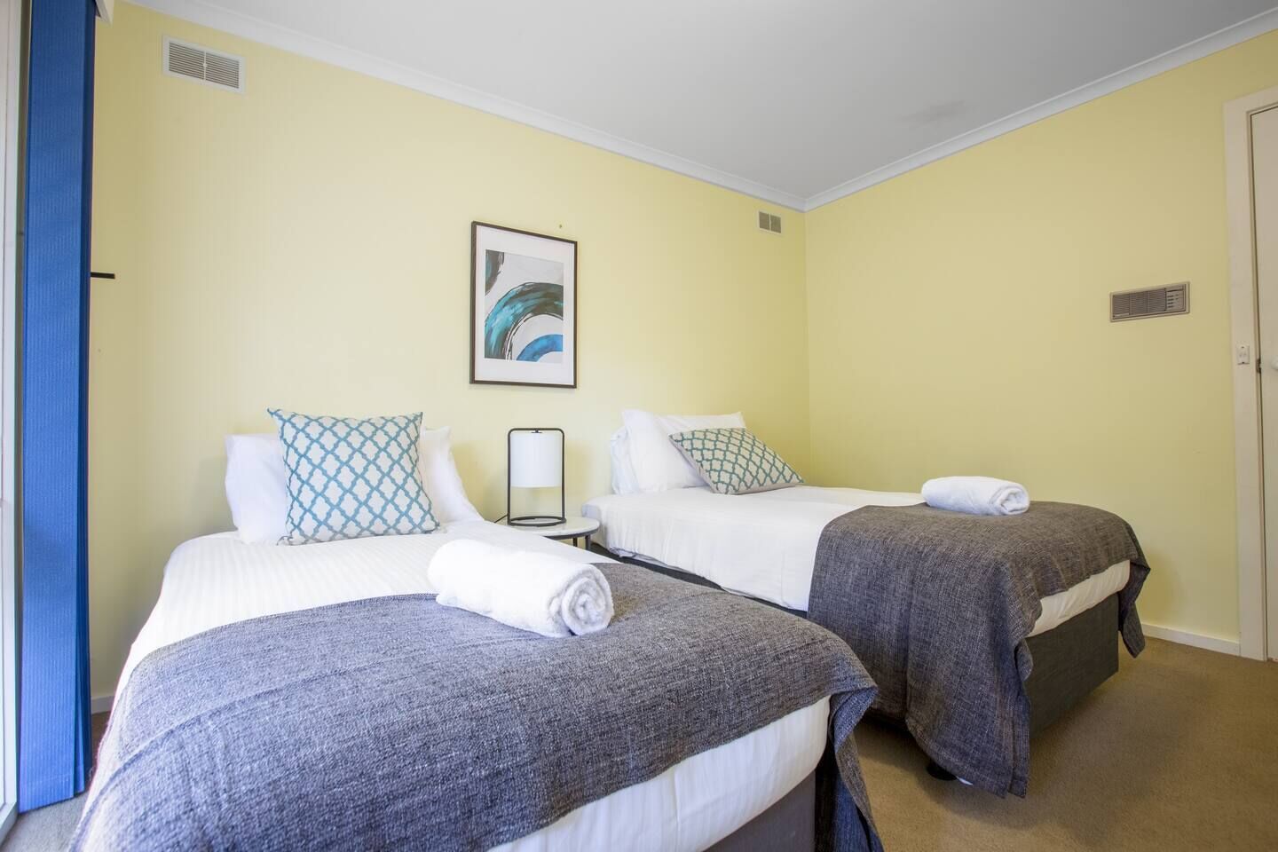 A hotel room with two beds, each with a grey blanket and white linens. There is a framed artwork on the wall above the beds, and a small table with a lamp between them.