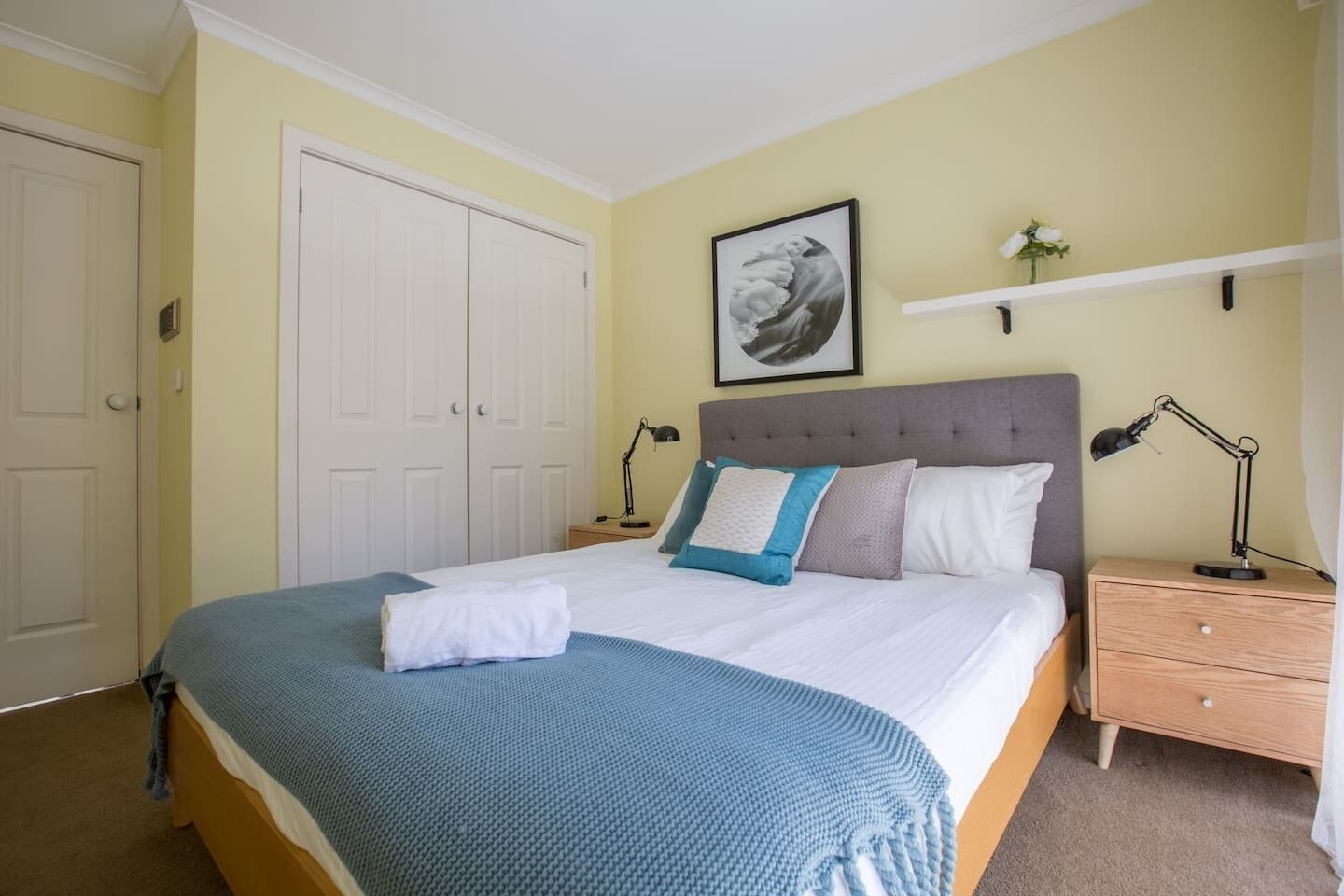 A neatly made bed with a blue blanket, a grey headboard, and a framed picture on the wall.