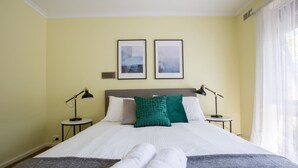 Ein Hotelzimmer mit einem großen Bett, zwei Nachttischen, zwei Lampen, einem Fenster mit durchsichtigen Vorhängen und zwei gerahmten Bildern an der Wand.