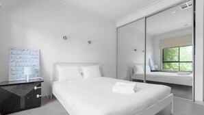 A modern bedroom with a large bed, a bedside table, a mirror, and a window with a view of greenery. - Spacious 3bedrooms big House@mitcham (Mitcham)
