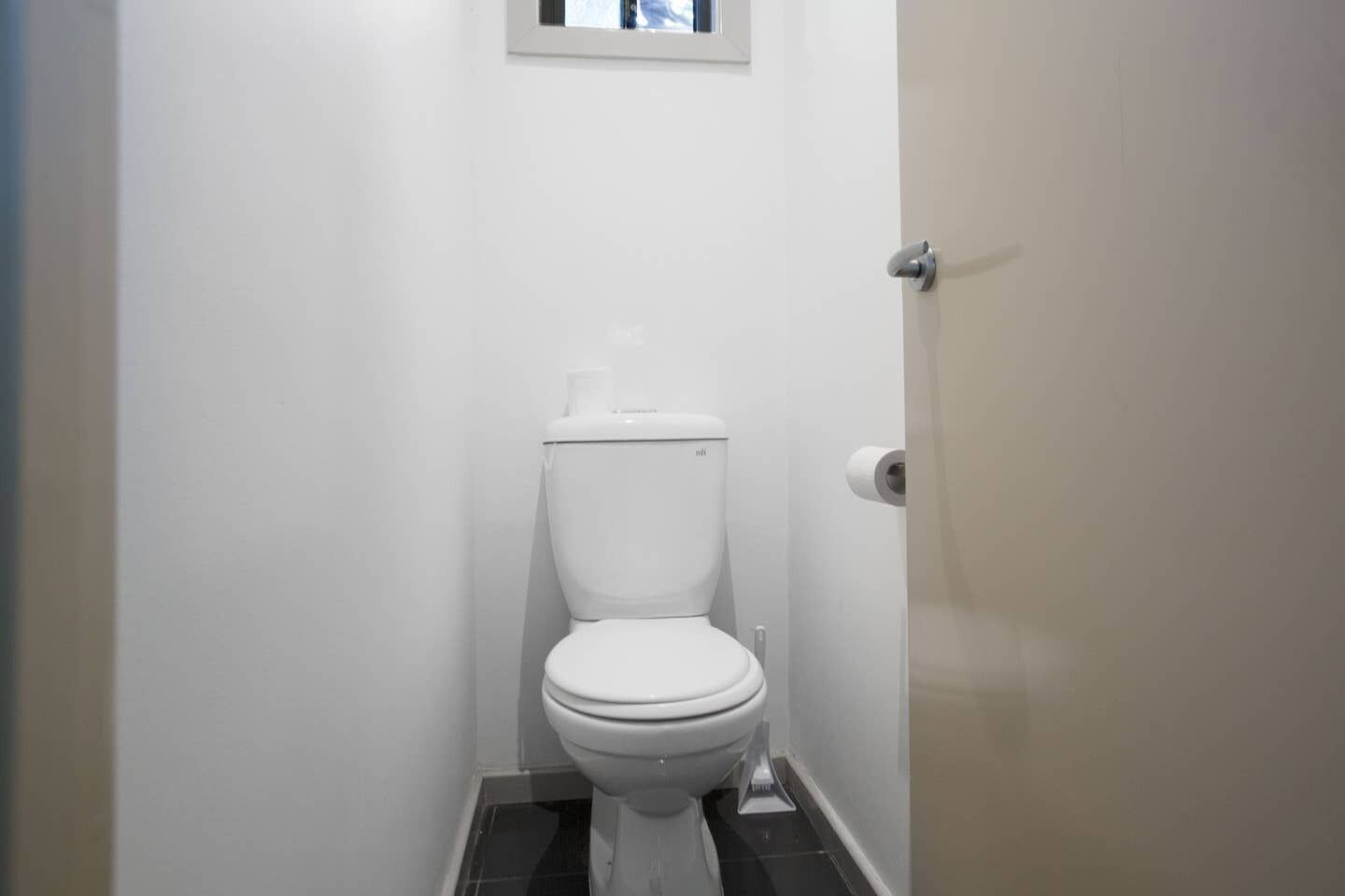 A small, white bathroom with a toilet, a small window above the toilet, and a door on the right.