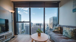 A modern living room with a large window, a flat-screen TV, a sofa, and a small table with a plant. - Modern Living@collins Centro Location 2BR (Melbourne)