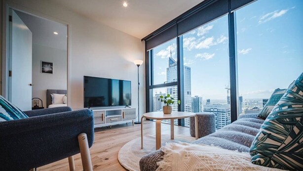 A modern living room with a large TV, a sofa, a coffee table, and a view of the city skyline. - Modern Living@collins Centro Location 2BR (Melbourne)