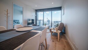 A modern living room with a dining area, large windows, and a view of the cityscape. - Modern Living@collins Centro Location 2BR (Melbourne)