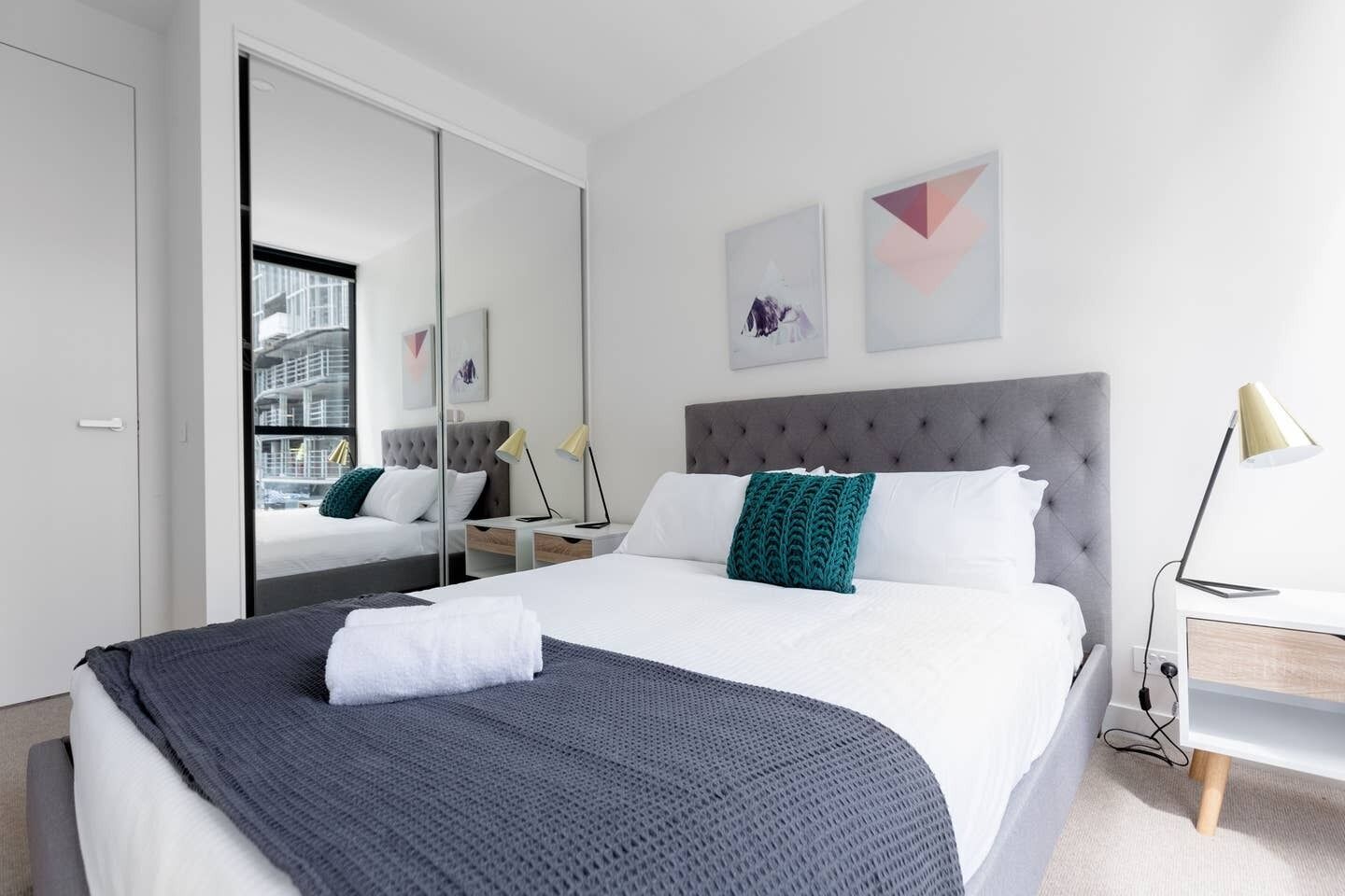 A modern hotel room with a large bed, a grey tufted headboard, a bedside table with a lamp, and a large mirror.