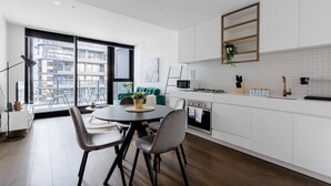 A modern kitchen with white cabinets, a dining area with a round table and chairs, and a view of the city through large windows. - Perfect Location 2 Bdrs Apartment@glen Waverley (Glen Waverley)