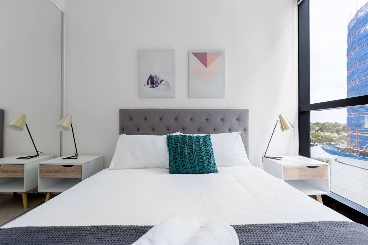 A modern hotel room with a large bed, two bedside tables with lamps, a grey tufted headboard, and artwork on the wall.