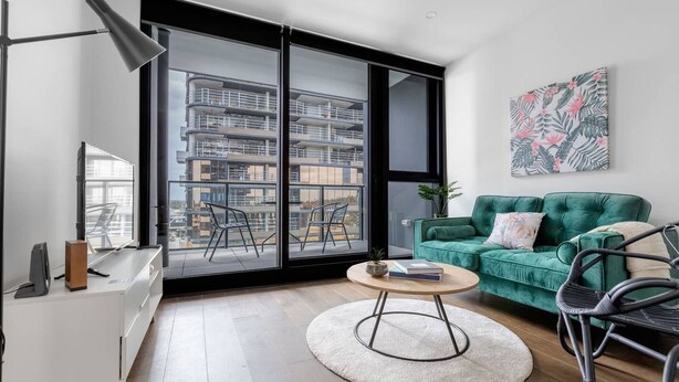 A modern living room with a green sofa, a round coffee table, and a view of a high-rise building through large windows. - Perfect Location 2 Bdrs Apartment@glen Waverley (Glen Waverley)