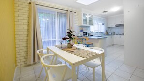 Una cocina luminosa con armarios blancos, un comedor con mesa y sillas, y un amplio ventanal con cortinas.