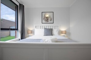 A neatly made bed with white bedding and a grey pillow, two bedside lamps, a framed artwork above the bed, and a window with curtains.