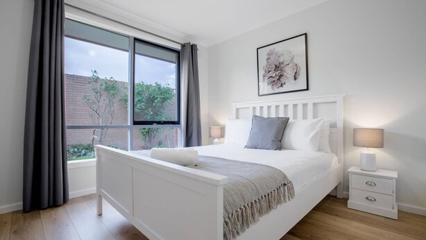 Un dormitorio moderno con cama blanca, cortinas grises, un ventanal grande y una obra de arte enmarcada en la pared.