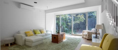 A modern living room with a sofa, armchairs, a coffee table, and a view of a garden through large sliding glass doors.