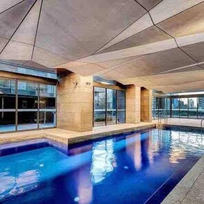A modern indoor swimming pool with clear glass walls, a tiled floor, and a concrete ceiling.