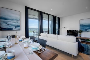 A modern living room with a dining area, large windows, a sofa, a TV, and artwork on the walls. - Breathtaking Lake/parkview Apt Pool&gym@3br (Melbourne)