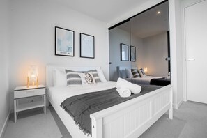 Un dormitorio moderno con una cama grande, una mesita de noche con lámpara, un espejo y dos cuadros abstractos en la pared.