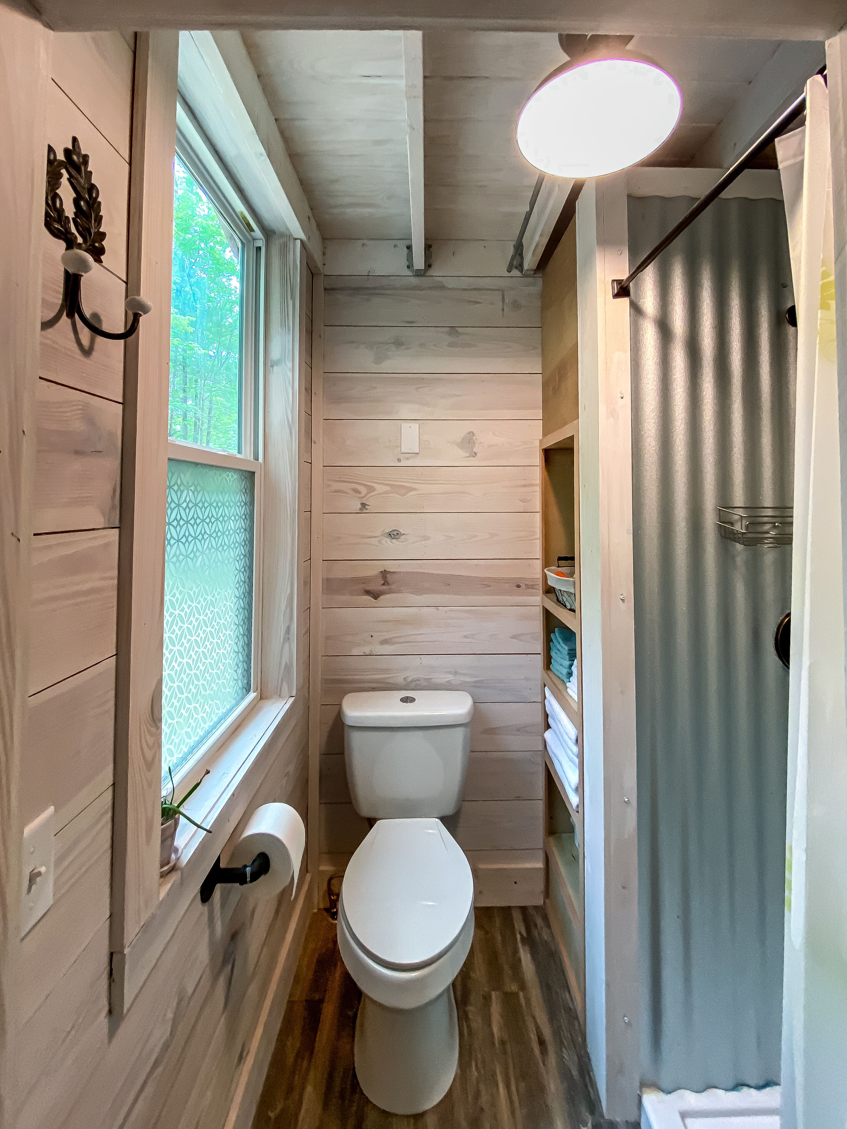 Ein kleines, schmales Badezimmer mit Holzwänden, einer Toilette, einem Fenster mit Milchglas und einer Duschvorhang.