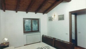A bedroom with a wooden ceiling, a bed with a striped pillow, a nightstand with a lamp, a window, and a radiator.
