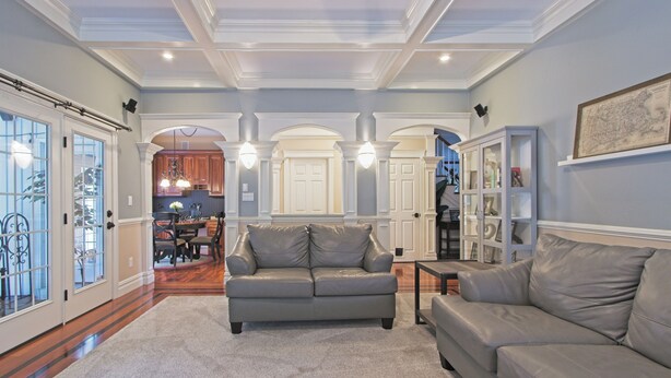 A spacious living room with a grey leather sofa, a coffee table, and a bookshelf. There is a dining area visible in the background with wooden cabinets and a chandelier. - Large Luxury Family Home Near Five Colleges. The TOP short term rental in area! (Hadley)