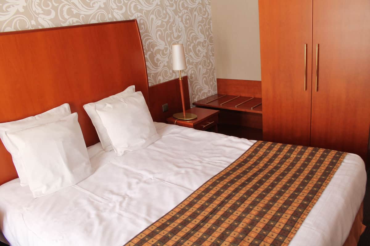 A hotel room with a wooden headboard, a bed with white linens, a patterned bedspread, a nightstand with a lamp, and a wardrobe.