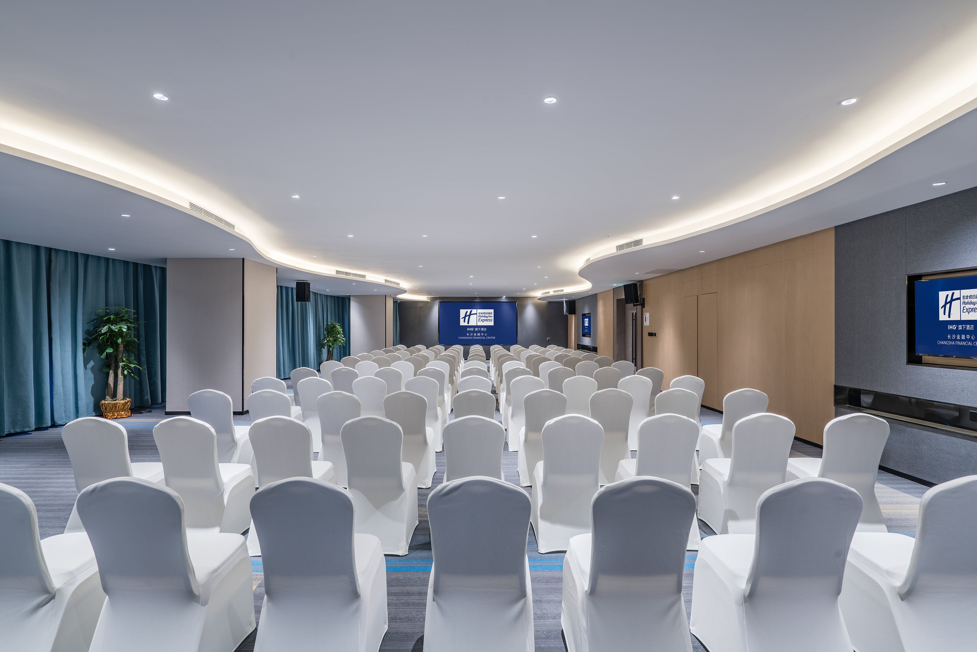 Un salón de conferencias con filas de sillas blancas, un escenario con un banderín azul y un diseño moderno en el techo.