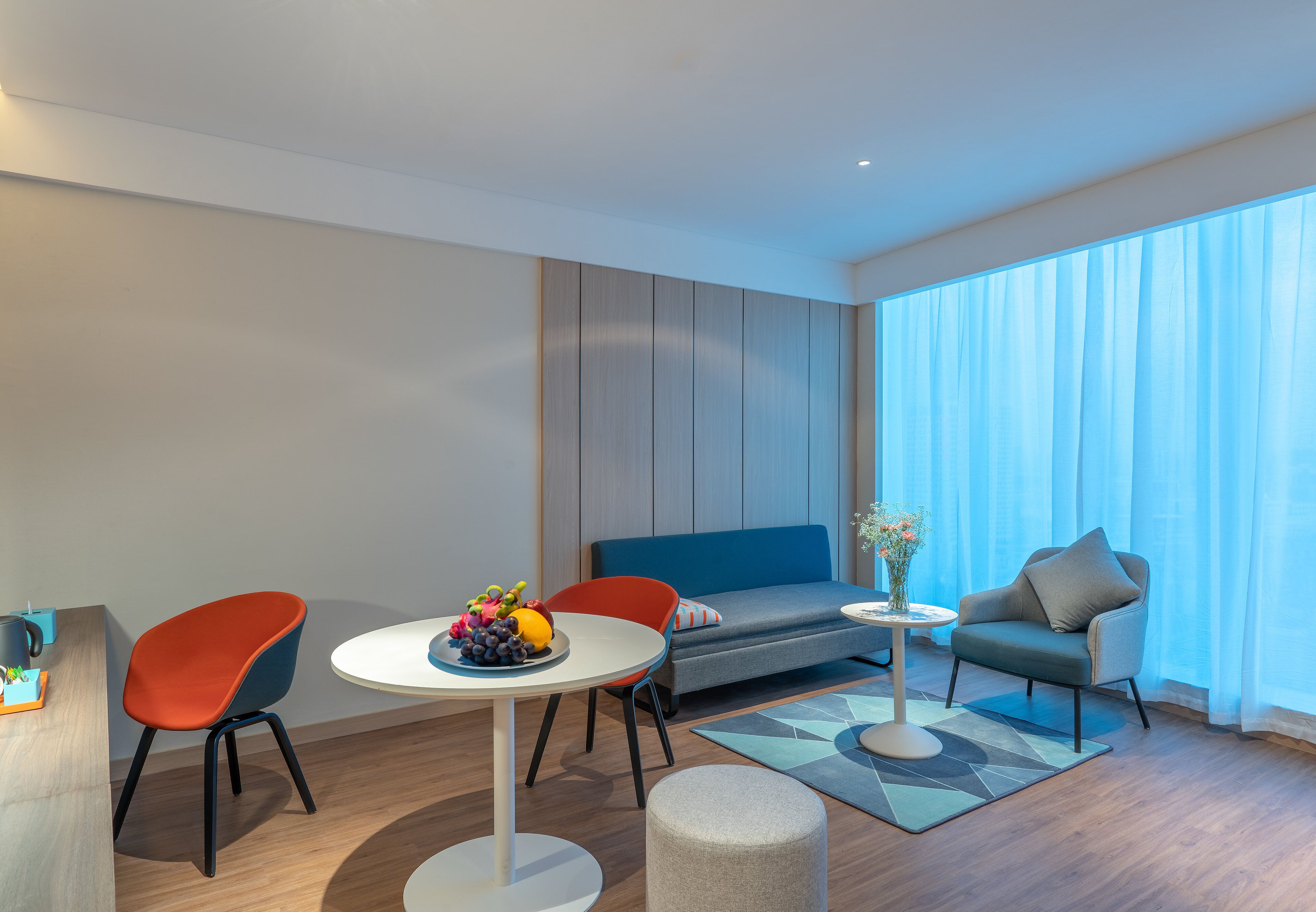 A modern living room with a sofa, chairs, a small table with fruit, and a large window with curtains.