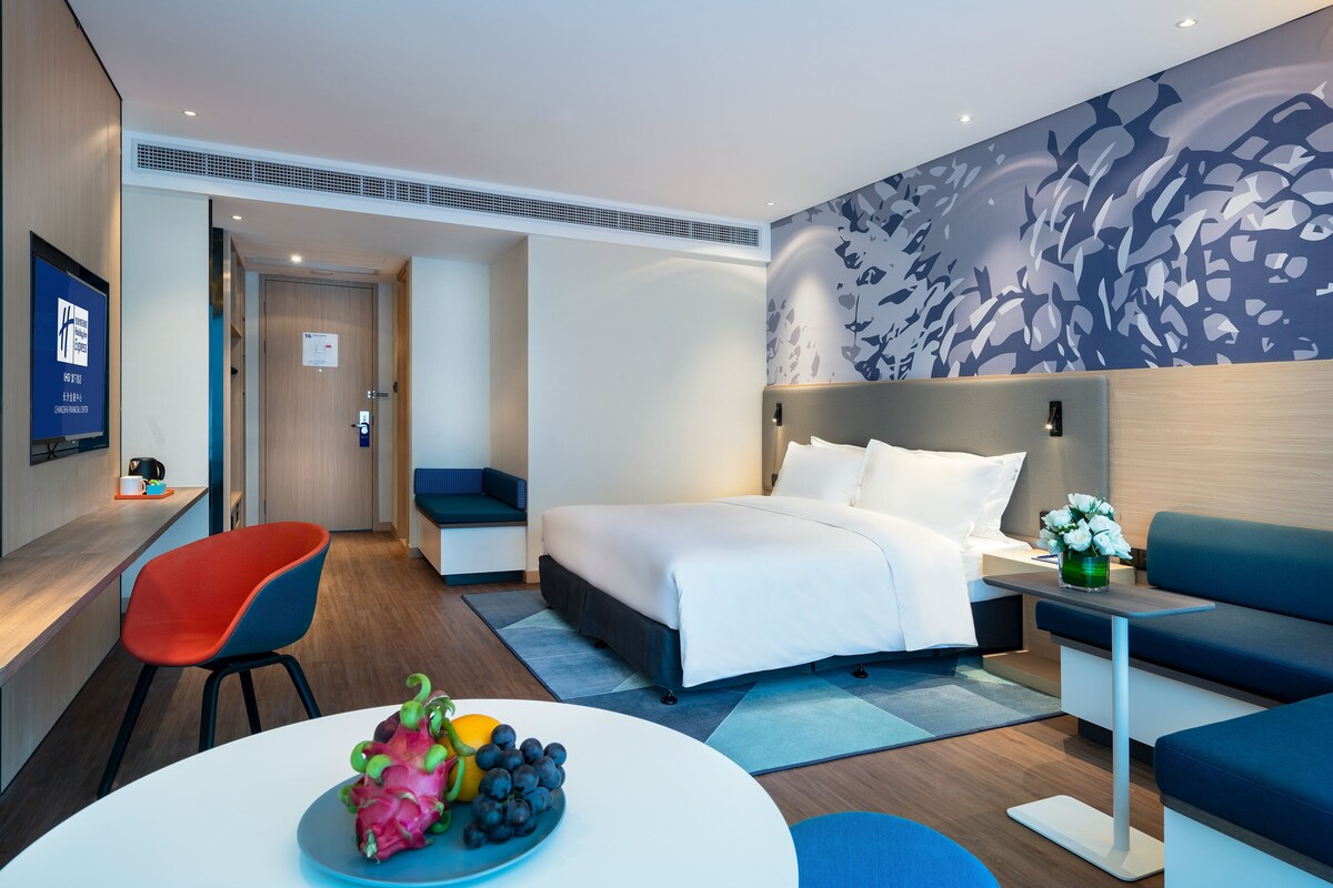 A hotel room with a bed, a desk, a chair, a table with fruit, and a wall with a leaf pattern.