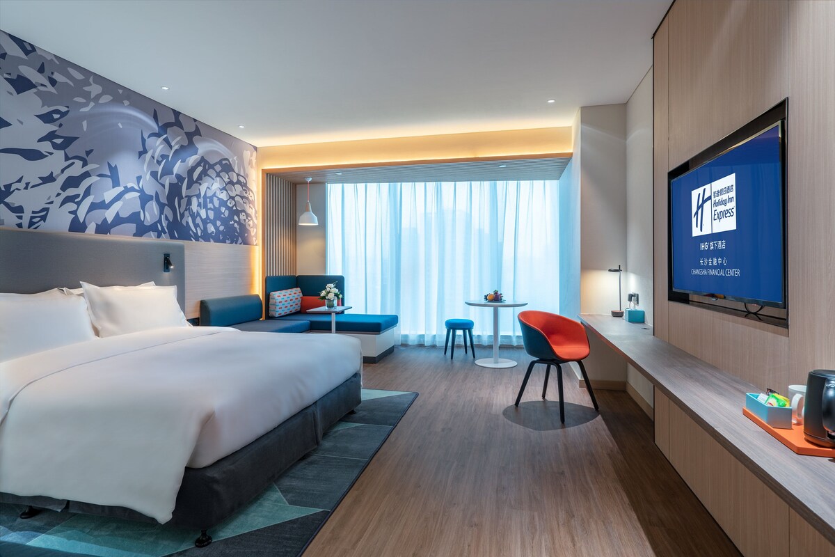 A hotel room with a bed, a seating area with a table and chairs, a television, and a large window with curtains.