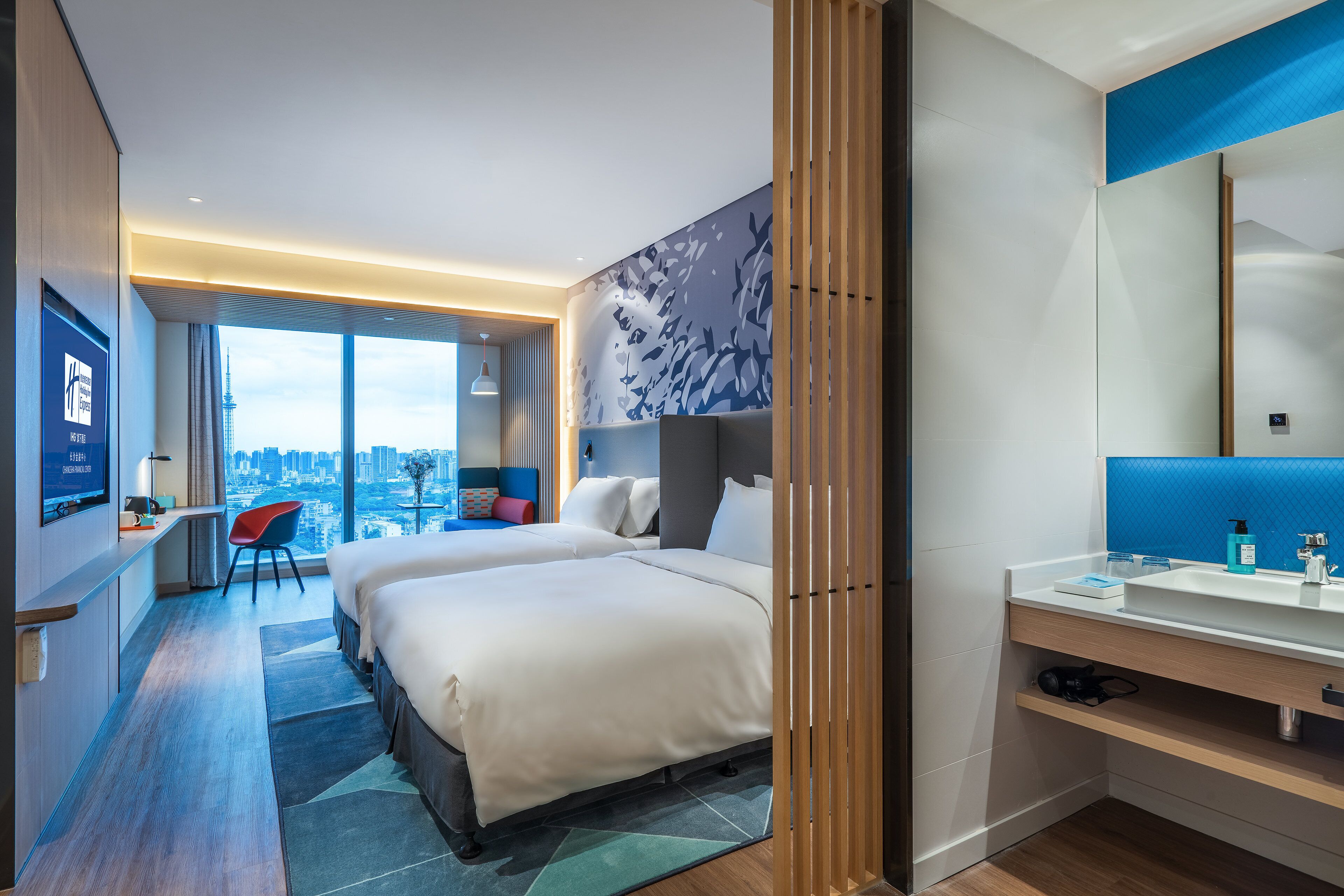 A modern hotel room with a large bed, a desk, a chair, a bathroom with a mirror, and a view of the city.