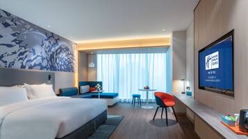 Una camera d'albergo moderna con un letto grande, una zona salotto con divano e poltrone, una TV a parete e vista sulla città da ampie finestre.