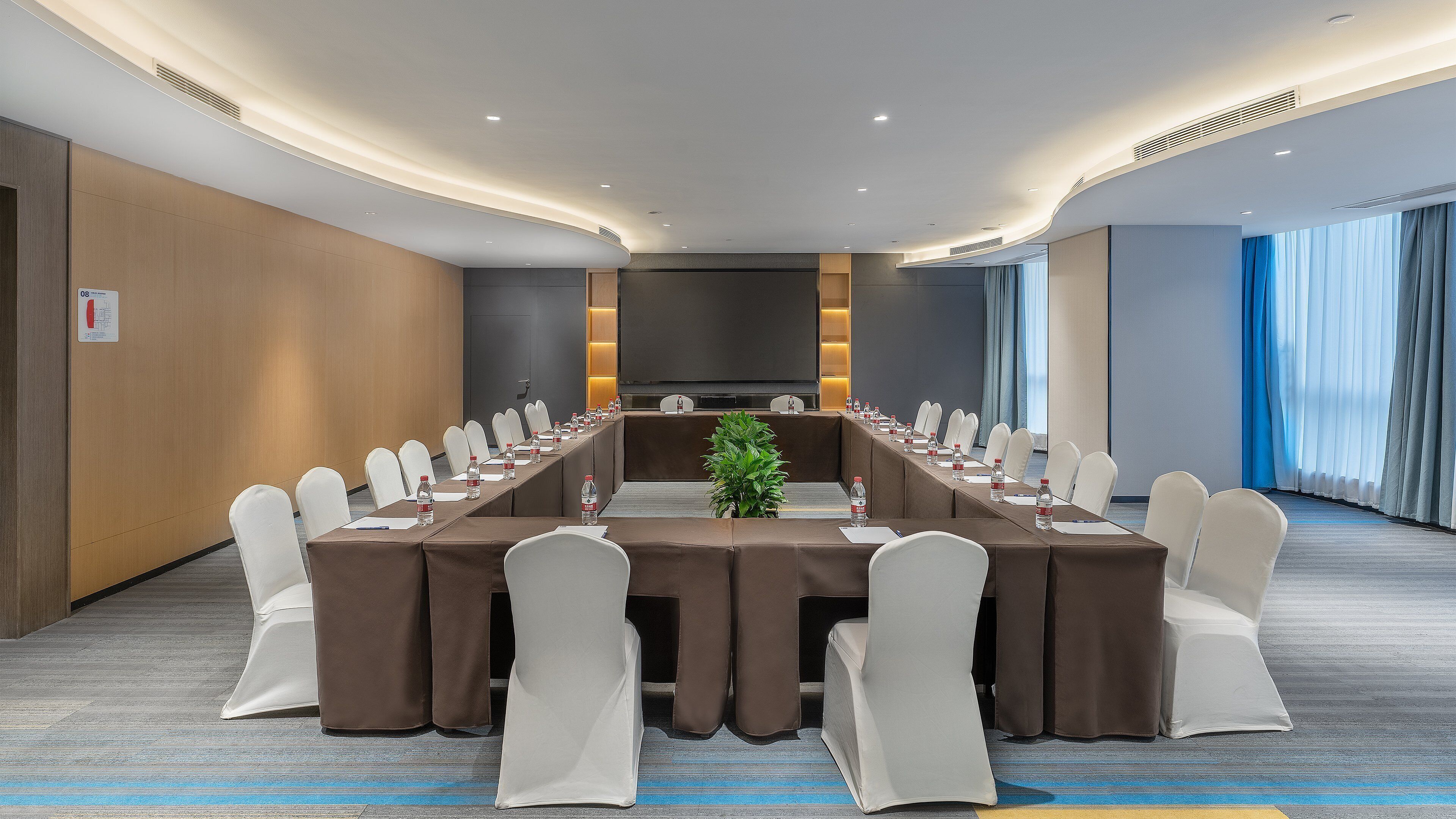 Un salón de conferencias con una larga mesa, sillas blancas y una pantalla grande.