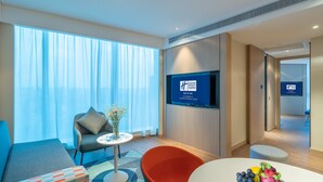Quarto de hotel moderno com uma janela ampla, TV de tela plana, sofá confortável, poltrona elegante e uma cesta de frutas sobre a mesa.
