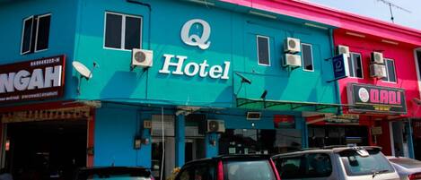 Un edificio de hotel con un llamativo letrero de 'Q Hotel', varios equipos de aire acondicionado y coches estacionados en la parte delantera.