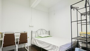 Una cama individual con estructura metálica, un escritorio con dos sillas y un estante metálico con ropa doblada.