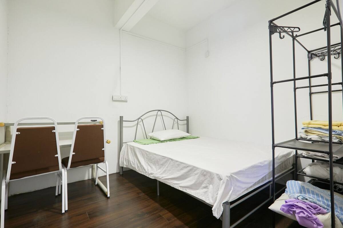A single bed with a metal frame, a desk with two chairs, and a metal shelf unit with folded clothes.