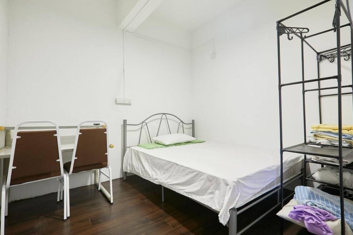 A small, single-bed room with a metal bed frame, a desk with two chairs, and a metal shelf unit with folded clothes.
