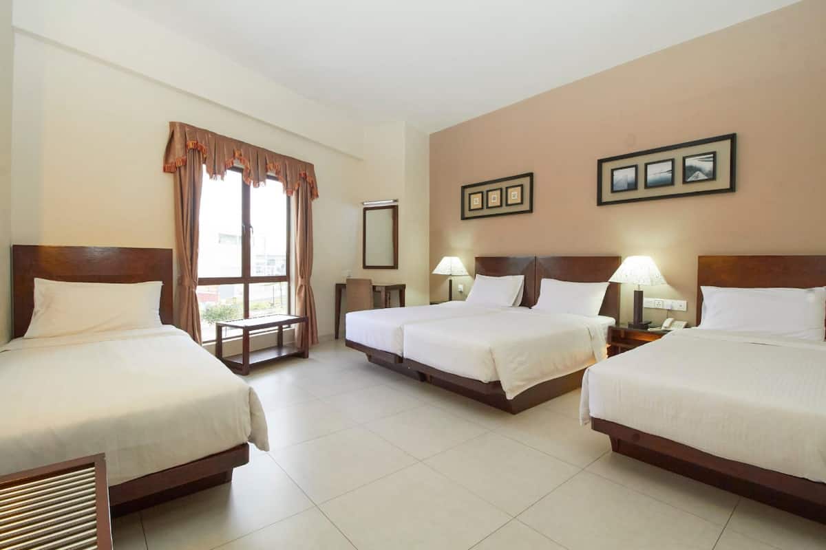 A hotel room with four single beds, a large window with curtains, a small table, a chair, and a wall-mounted mirror.