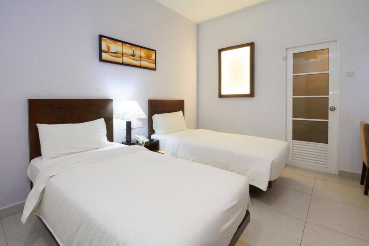 A hotel room with two beds, a nightstand with a lamp, a painting on the wall, and a sliding door closet.