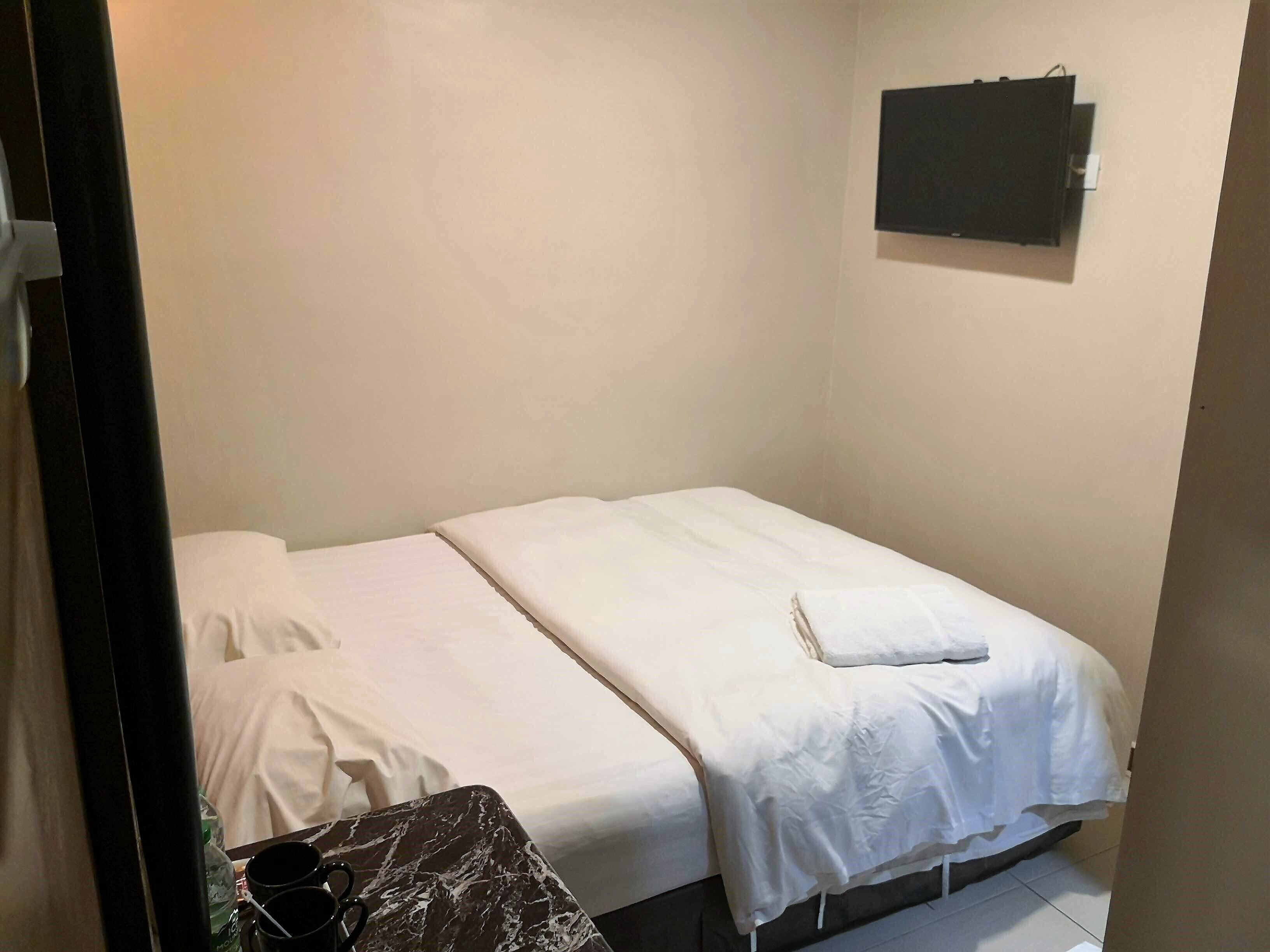 Une petite chambre avec un lit simple, recouverte d’une courtepointe blanche unie, une petite table avec une nappe à motifs de marbre et un tableau noir accroché au mur.