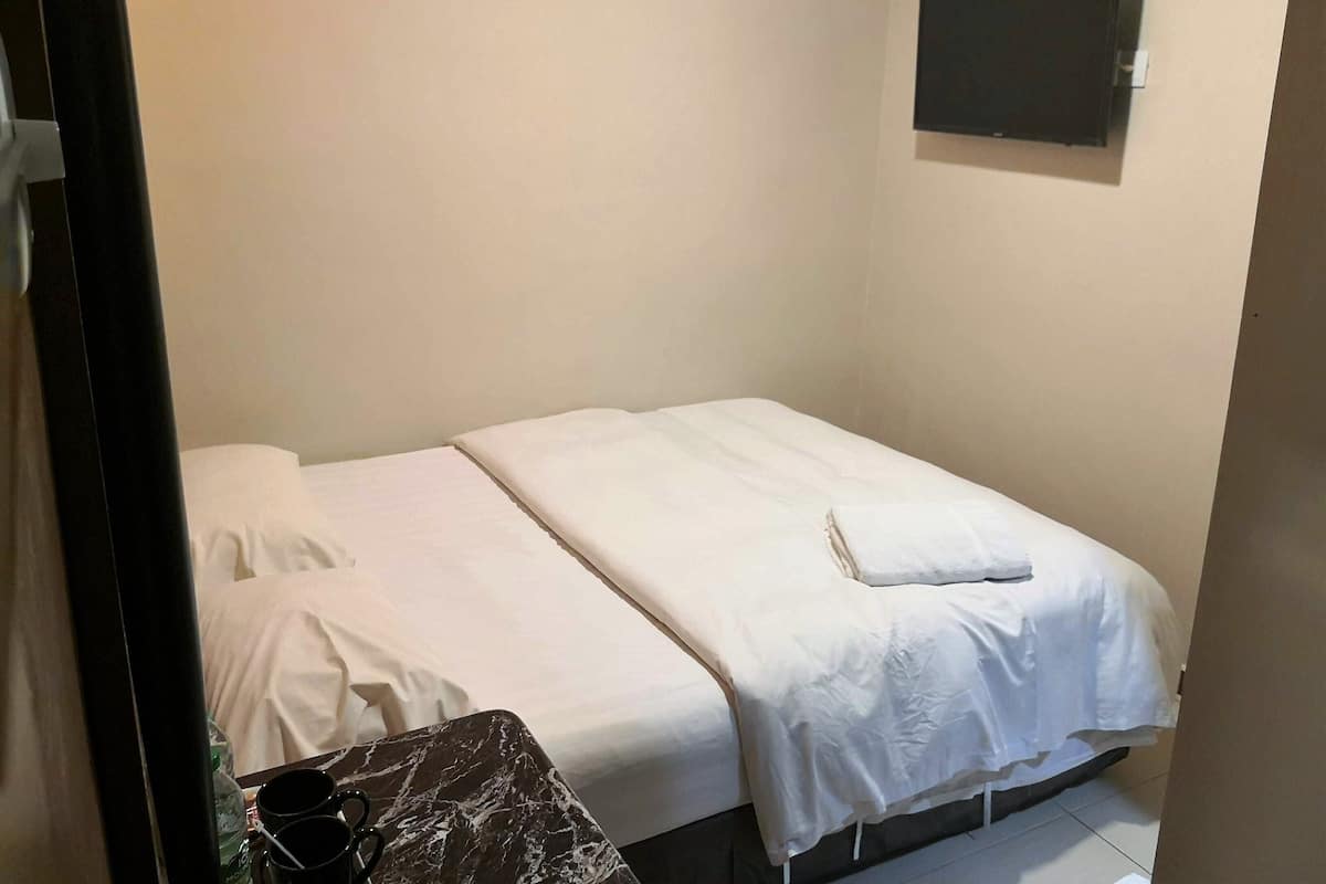 Une petite chambre avec un lit simple, recouverte d’une courtepointe blanche unie, une petite table avec une nappe à motifs de marbre et un tableau noir accroché au mur.