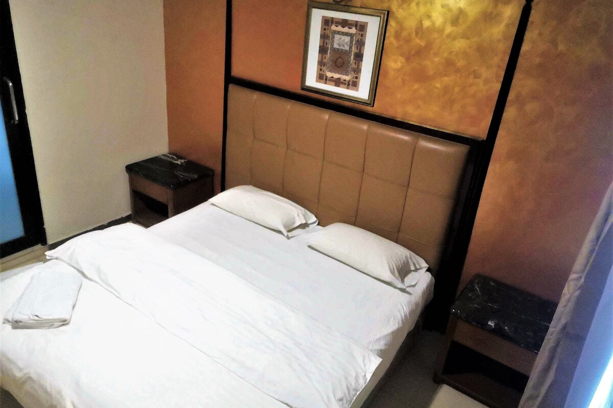 A hotel room with a bed, a nightstand, a framed picture on the wall, and a wall-mounted light fixture.