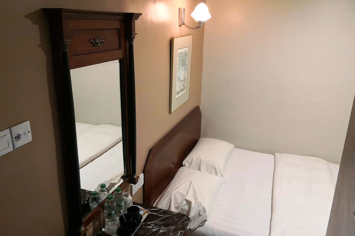 A hotel room with a bed, a bedside table with a lamp, a mirror, and a wall-mounted picture.