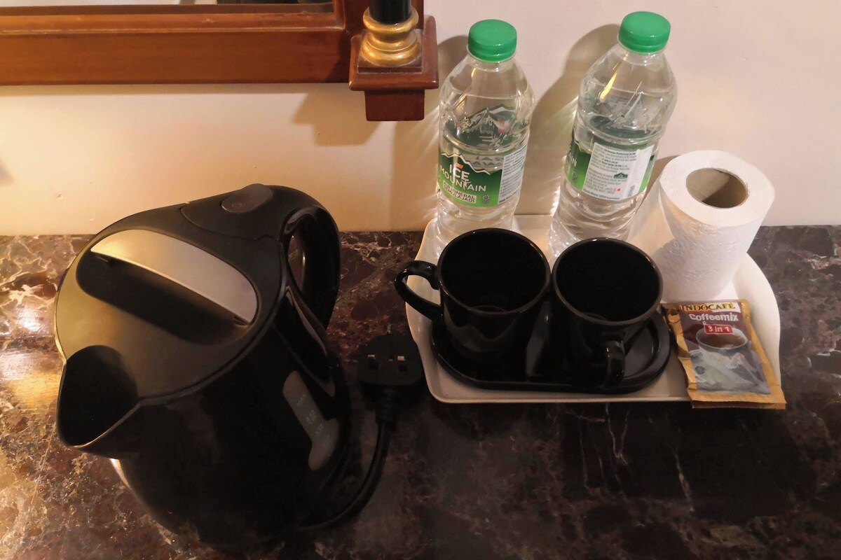 A coffee maker, two black mugs, two bottles of water, a roll of toilet paper, and a packet of coffee mix on a marble countertop.