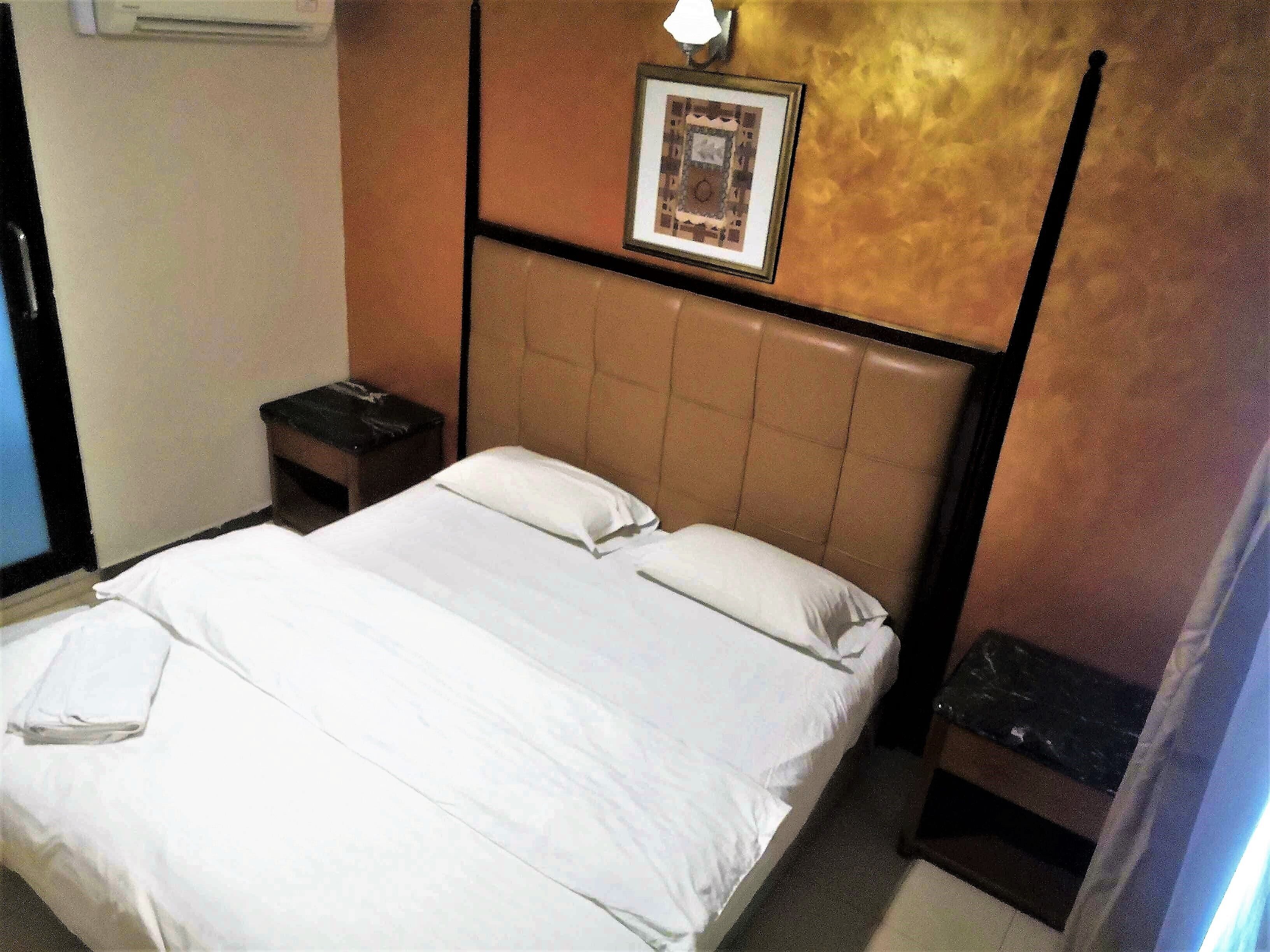 Une chambre d’hôtel avec un lit, une table de chevet, un tableau encadré au mur et un luminaire fixé au mur.
