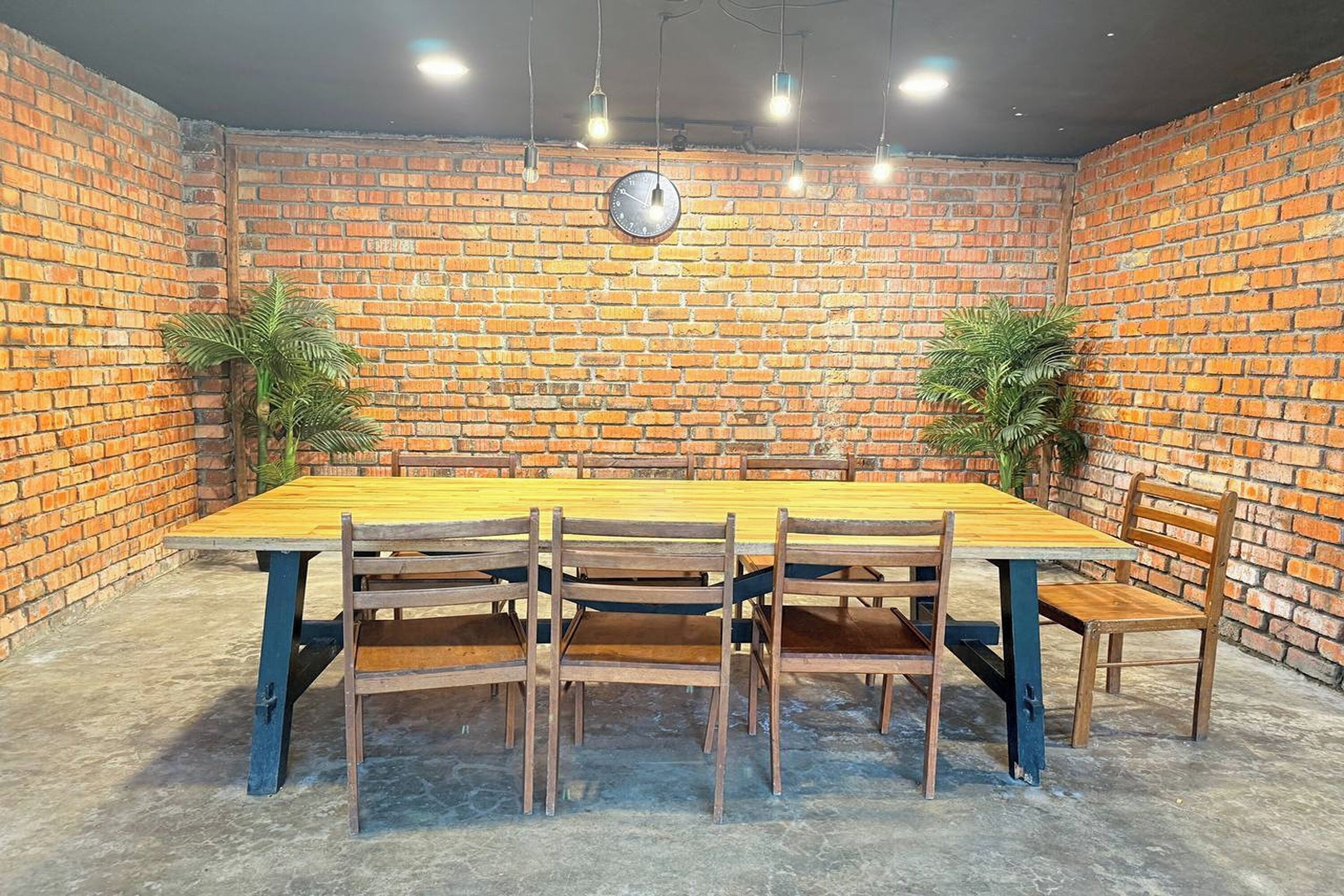 A brick-walled room with a wooden table and chairs, a clock on the wall, and hanging lights.