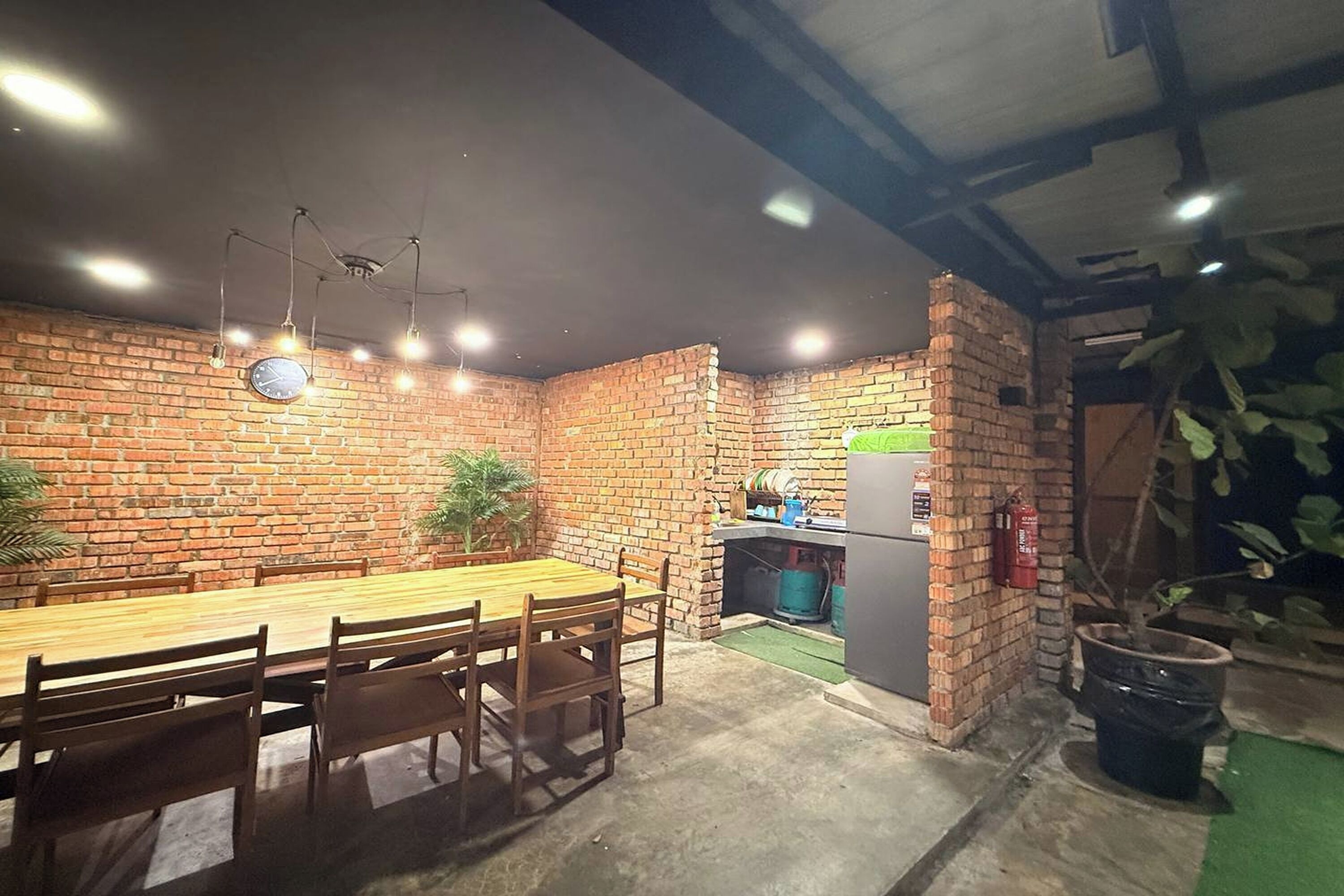 A dining area with a brick wall, wooden table, and chairs. There is a small kitchen area with a refrigerator and a fire extinguisher.
